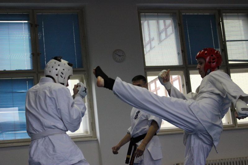Dva závodníci v bílých kimonech a ochranných přilbách při zápasu v karate v tělocvičně.